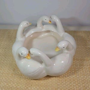 May include: A white ceramic bowl featuring five sculpted goose figures arranged around the perimeter. Each goose has a smooth, glossy finish with a small yellow beak and black eyes, forming a circular basin in the center of the vessel.
