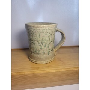 May include: A handmade ceramic mug with a textured surface and a light green glaze. The mug has a handle and a wide base, and it is approximately 4 inches tall. The mug is sitting on a wooden surface.