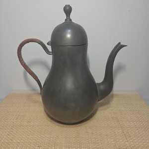 May include: A matte gray metal teapot with a rounded body, a curved spout, and a lid featuring a small knob handle. The side handle is wrapped in a textured brown material, providing a contrast to the smooth, dark metallic finish of the vessel.