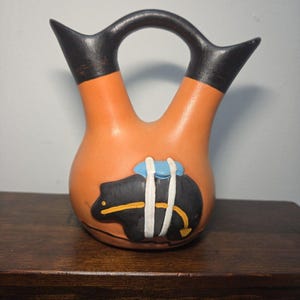 May include: A terracotta-colored ceramic vessel with two spouts connected by a bridge handle. The vessel features a black bear graphic with a blue and white pack on its back, and the upper portions of both spouts are painted in matte black.