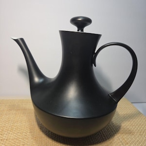 May include: A matte black teapot with a wide, rounded base, a slender curved spout, and a tall, tapered neck. The teapot features a matching black lid with a small, rounded knob handle and a thin, curved handle on the side for pouring.