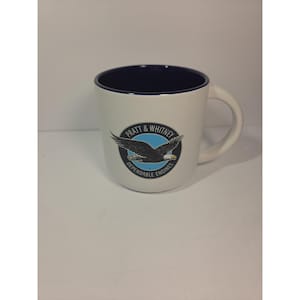 May include: A white ceramic mug with a dark blue interior and handle. The front features a circular logo with a light blue background, a black eagle graphic, and the text Pratt & Whitney Dependable Engines printed in black around the border.