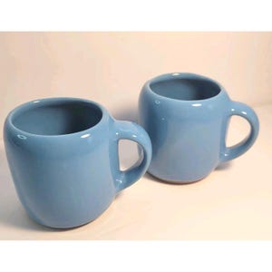 May include: Two light blue ceramic mugs with rounded bodies and large, curved handles. The mugs have a glossy finish and are set against a plain background. The mugs are designed for beverages.
