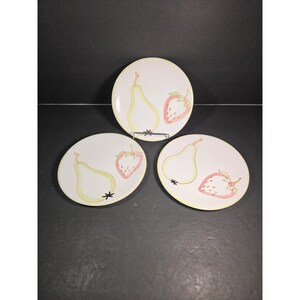 May include: Three white ceramic plates with a fruit design. Each plate features a yellow pear and a red strawberry illustration. The plates have a yellow border and black star accents. The plates are arranged on a dark surface.