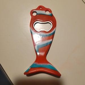 May include: A red, white, and blue fish-shaped bottle opener. The opener has a metal opening and a hole for hanging. The design features stripes of red, white, and blue, creating a unique and decorative kitchen tool.