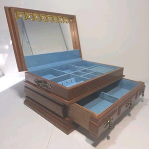 May include: A wooden jewelry box with a hinged lid featuring an interior mirror and gold-tone necklace hooks. The box has a light blue velvet-lined interior with multiple compartments, ring slots, and a pull-out drawer with ornate metal handles.