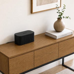 Puede incluir: Una caja organizadora de cables rectangular en color negro mate con una textura acanalada vertical en sus lados exteriores y una ranura horizontal estrecha en la parte superior para el paso de cables, diseñada para ocultar regletas y cables.