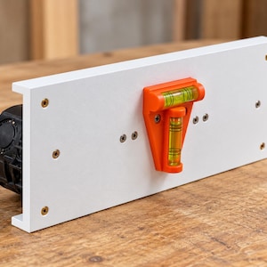 Könnte beinhalten: Eine weiße rechteckige Montageplatte mit einer leuchtend orangefarbenen Wasserwaage mit zwei Libellen in der Mitte. Die Platte ist mit kleinen Metallschrauben befestigt und mit einem schwarzen runden Bauteil auf der linken Seite verbunden.