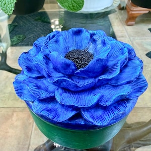 May include: A vibrant blue ceramic flower sculpture with a dark center. The flower has multiple layers of petals and sits on a green base. The sculpture is a decorative piece.