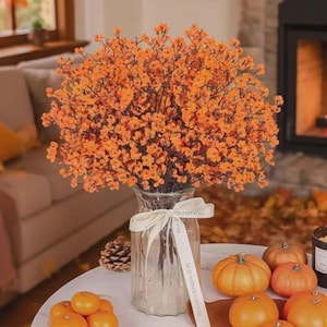 May include: A large bouquet of vibrant orange baby's breath flowers arranged in a clear glass vase. The vase is tied with a white ribbon featuring printed text. The arrangement is displayed on a table alongside several small orange pumpkins.