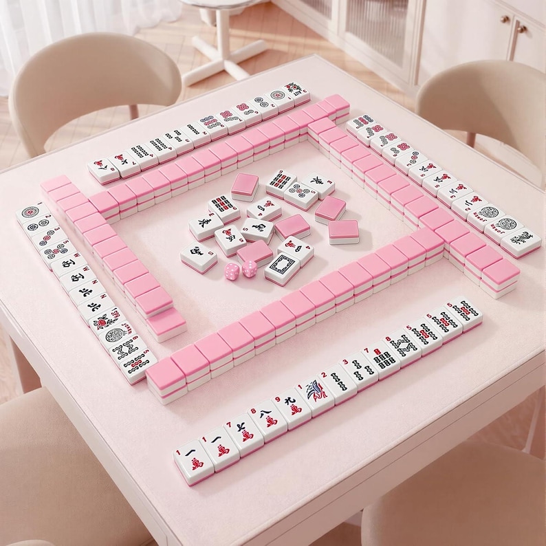 May include: A complete Mahjong set arranged on a light pink table. The tiles are white with red and black symbols, and pink edges. The set includes racks and loose tiles, ready for play. The table is surrounded by chairs.