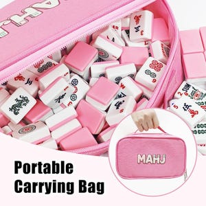 May include: A pink portable carrying bag filled with white and pink Mahjong tiles. The bag is open, revealing the tiles inside. The bag has a zipper and a handle. The text "MAHJ" is on the bag.