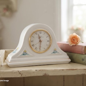 May include: A white ceramic mantel clock with an arched top and a gold-rimmed circular face featuring Roman numerals. The clock displays the text Laura Ashley and includes small floral motifs with yellow and blue accents on the base.