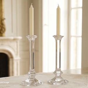 May include: A pair of clear glass candlesticks with a faceted, multi-tiered base and a slender stem, each holding a single cream-colored taper candle. The design features a flared top rim and a geometric, stepped pedestal base for stability.