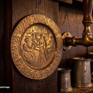 May include: A circular brass decorative plate featuring an embossed scene of figures gathered around a table, mounted on a dark wood panel. Beside it, a brass beer tap extends over several metallic tankards resting on a wooden surface.