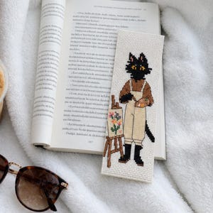 May include: A rectangular cross-stitch bookmark featuring a black cat wearing tan overalls and holding a paint palette, standing next to an easel with a floral painting. The bookmark rests on an open book next to iced coffee, a cookie, and sunglasses.