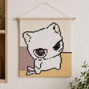 May include: A square crochet wall hanging featuring a pixelated white cat design. The cat has large dark eyes and is set against a background of cream, muted mauve, and mustard yellow blocks. It hangs from a wooden dowel with a white cord.