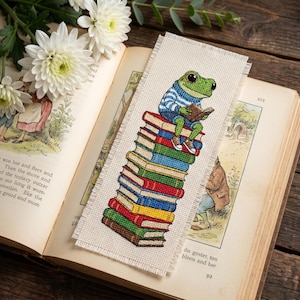 May include: A handmade bookmark featuring a cross-stitched frog reading a book while sitting atop a stack of colorful books. The bookmark is made of natural-colored fabric and is placed inside an open book with illustrations and text. White flowers are in the background.