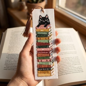 Könnte beinhalten: Handgefertigtes Lesezeichen mit einer schwarzen Katze auf einem Stapel bunter Bücher. Das Lesezeichen besteht aus weißem Stoff mit Kreuzstichstickerei. Oben hängt eine braune Quaste. Die Bücher sind in verschiedenen Farben.