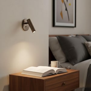 Peut inclure: Une liseuse murale en bronze foncé dotée d'une tête cylindrique et d'une plaque de base circulaire. Le luminaire comporte un petit interrupteur à bascule sur la base et projette un faisceau de lumière chaude sur un livre ouvert posé sur une table.