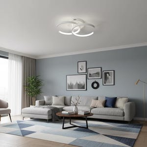 Puede incluir: Un salón moderno con una lámpara de techo LED blanca con tres anillos circulares que se cruzan. La habitación incluye un sofá seccional gris, una mesa de centro redonda y una alfombra con estampado geométrico en tonos azules y grises. Obras de arte adornan la pared azul claro.