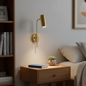 May include: A gold-toned wall-mounted lamp featuring a cylindrical head and a slender vertical arm attached to a circular base. The lamp is illuminated, casting a focused downward light, and includes a small black power switch on the base.