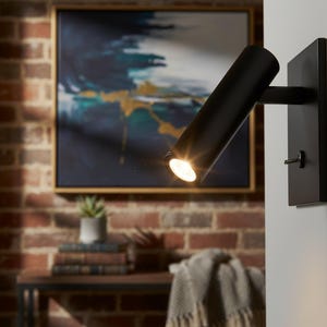 Könnte beinhalten: Ein schwarzer, an der Wand montierter Strahler mit zylindrischem Leuchtkopf und rechteckiger Basis. Das Licht ist eingeschaltet und beleuchtet die Umgebung. Die Leuchte ist an einer weißen Wand montiert, mit einer Backsteinmauer und Kunst im Hintergrund.