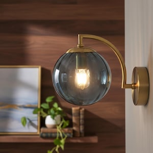May include: A modern wall sconce with a clear glass globe shade. The fixture has a brass finish and a curved arm. The light bulb inside the globe is illuminated, casting a warm glow. The sconce is mounted on a white wall.