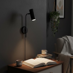 May include: A matte black wall-mounted lamp with a cylindrical head and a thin adjustable arm. The lamp is attached to a circular base plate and features a downward-facing light source that illuminates an open book on a wooden surface.