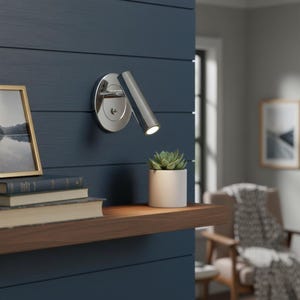 Peut inclure: Une applique murale spot en chrome poli avec une tête cylindrique et une plaque de base circulaire. La lumière est dirigée vers le bas, avec un petit interrupteur à bascule sur la base et une ampoule LED brillante visible au bout du tube.