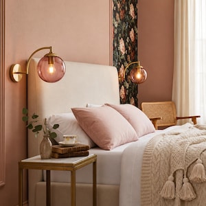 May include: Two wall-mounted lamps featuring brushed brass arms and spherical, translucent pink glass shades. Each lamp holds a single glowing light bulb, providing warm illumination to the bedroom space with a soft, diffused light effect.