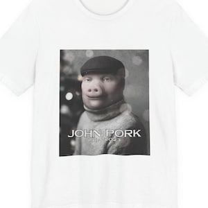 May include: A white t-shirt featuring a central graphic print of a pig wearing a dark flat cap and a turtleneck jumper. The text JOHN PORK is printed in white capital letters above the years 1984-2023 in a smaller white font underneath the image.