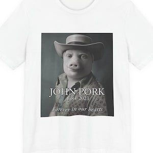 May include: A white short-sleeved t-shirt featuring a black and white graphic print of a pig wearing a cowboy hat and a patterned shirt. The text reads JOHN PORK 1984-2023 Forever in our hearts, centred below the pig illustration on the front.