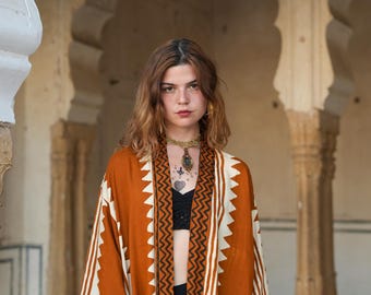 Puede incluir: Un kimono de color óxido con patrones geométricos blancos y negros. El kimono tiene un diseño en zigzag en los bordes y rayas verticales. La modelo lleva un top negro y joyas.
