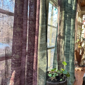 Puede incluir: Cortinas transparentes para ventanas en tonos morados y verdes con estampados florales. Las cortinas cuelgan frente a una ventana, con la luz del sol entrando. Una planta en maceta está frente a las cortinas.
