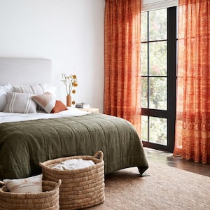 Puede incluir: Interior de dormitorio con una cama cubierta con una colcha verde acolchada, cojines y cestas tejidas. La habitación está decorada con cortinas naranjas estampadas y una alfombra de yute, creando un ambiente acogedor.