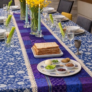 May include: A table set for a holiday meal, featuring a blue floral tablecloth and a purple and blue runner. The table is set with plates, glasses, and celery stalks. A plate of matzah and a Seder plate are visible, along with yellow flowers and lit candles.