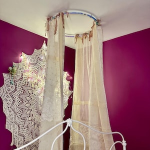 May include: A white metal bed frame with sheer, cream-colored curtains hanging from a circular canopy. The curtains are tied with brown ribbons. A large, white lace doily is on the wall, lit with fairy lights, against a magenta wall.