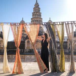 May include: A woman in a black dress stands between flowing curtains in front of a historic building. The curtains are various shades of beige, orange, and green. The building has a tall tower with multiple levels and intricate details.