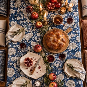 May include: A blue and white floral patterned tablecloth set for a meal with a braided loaf of bread on a wooden board, pomegranates, apples, ceramic plates with linen napkins, wine glasses, honey, and lit candles in brass holders.
