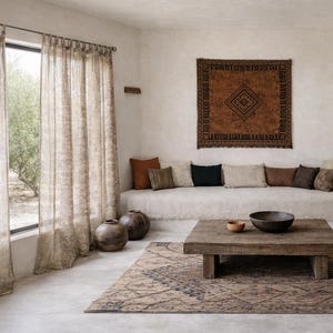 Puede incluir: Salón de tonos neutros con cortinas estampadas, sofá bajo y mesa de centro de madera. Una alfombra estampada y un tapiz de pared añaden textura. El espacio está decorado con cojines y objetos decorativos.