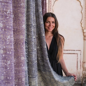 May include: A woman with long brown hair smiles while standing behind a patterned curtain. The curtain is a light gray with a floral design and a darker gray border. The woman is wearing a black sleeveless dress.