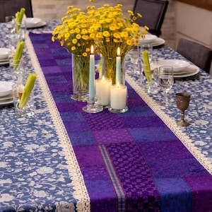 May include: A dining table set for a meal, featuring a blue floral tablecloth and a purple patchwork runner with a cream lace trim. Yellow flowers in glass vases, lit candles, and place settings complete the elegant arrangement.
