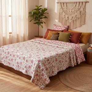 May include: A bed is covered with a white quilt featuring a pink floral pattern. The bed is adorned with various pillows in shades of pink, green, and gold. A macrame wall hanging and a potted plant add to the room's decor.