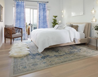 Puede incluir: Dormitorio con una paleta de colores neutros. La habitación cuenta con una cama con ropa de cama blanca, una alfombra estampada, cortinas azules, una silla de madera y una alfombra de piel sintética. Luces colgantes decorativas y una mesita auxiliar completan el espacio.