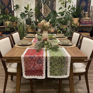 May include: A wooden dining table set for a meal, featuring a red and green patterned table runner with white lace trim. The table is set with neutral-toned plates and bowls, surrounded by chairs with cream-colored upholstery. The room is decorated with plants and tapestries.