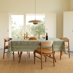 Op de afbeelding: Een eethoek met een rechthoekige tafel bedekt met een groene bloemen tafelkleed. De tafel wordt omringd door houten stoelen met beige zittingen. Een fles wijn, brood en kaarsen staan op de tafel.