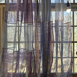 May include: A pair of sheer curtains with a light blue and purple pattern. The curtains are hanging in a window and are letting in natural light.