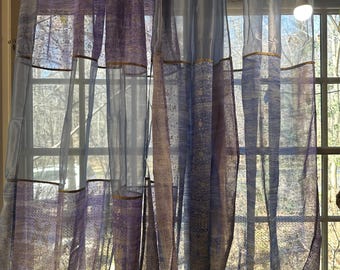May include: A pair of sheer curtains with a light blue and purple pattern. The curtains are hanging in a window and are letting in natural light.
