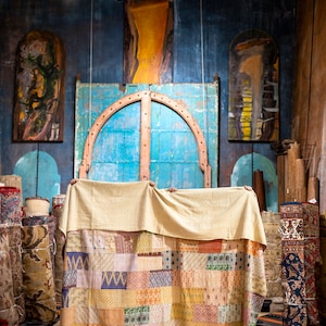 May include: A large patchwork quilt with a variety of colors and patterns, including pink, yellow, blue, and green. The quilt is being held up by two people, and it is covering a doorway in a room with a blue wall and several paintings.